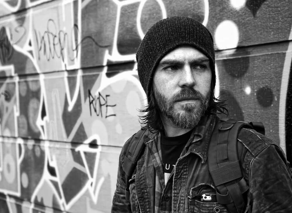 Person standing in front of graffiti wall with a knitted hat and backpack, looking away contemplatively in urban setting.