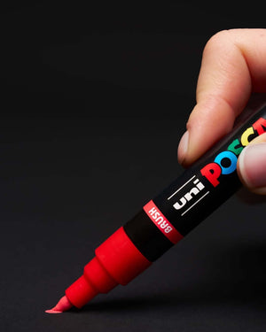 Hand holding red POSCA Brush PC-5BR paint pen with semi-flexible tip for versatile strokes.