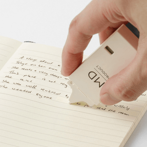 Hand using cream-colored correction tape on lined notebook page to conceal handwritten text.