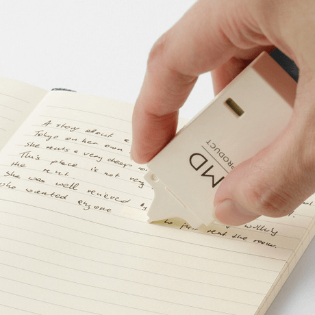 Hand using cream-colored correction tape on lined notebook page to conceal handwritten text.
