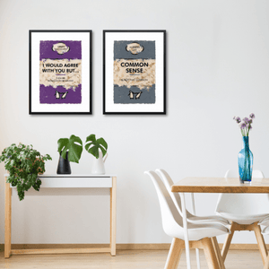 Modern dining room with two framed artworks on wall, 'I Would Agree With You But...' and 'Common Sense', white chairs and plants nearby.